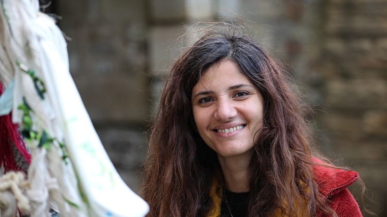Head and shoulders shot rtist Maryam Samaam smiling at the camera. She has long brown hair and is wearing a red coat