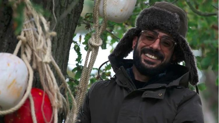 Photograph of Mike Shnsho smiling at the camera. He is wearing a woolly hat and standing beside a tree with the rope attached to it