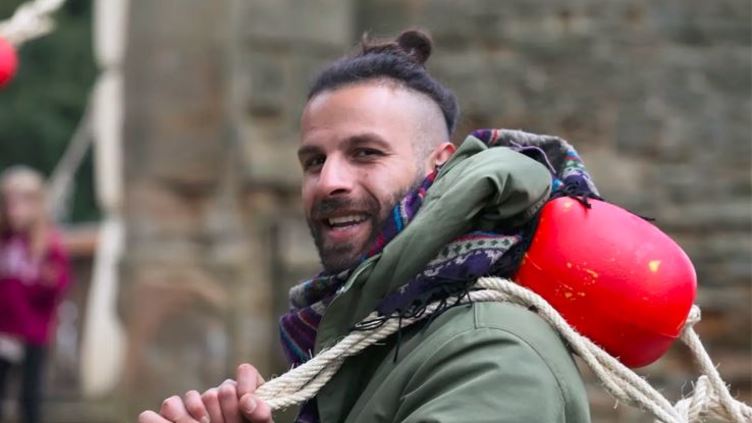 Photograph of Khaled Al Warea. He is holding a rope over his shoulder and smiling at the camera, wearing a green coat
