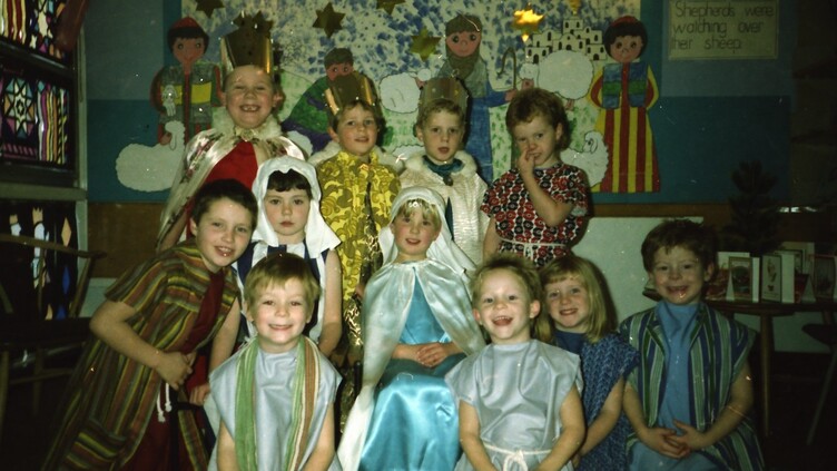 A group of young children in colorful costumes pose and smile for a photo, likely during a school nativity or Christmas play, with festive decorations and a mural in the background.