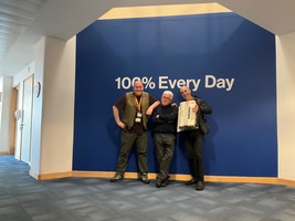 Three men posing in front of a wall with the sign '100% every day'