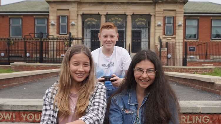 Three young people with game controllers sit on tiled steps outside a building