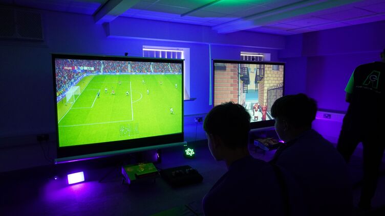 Two people play video games on large screens in a room lit with purple and green lights