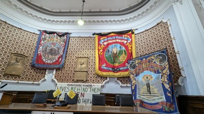 An ornate room with three large, colorful miners’ union banners hanging above a wooden table. The banners feature detailed illustrations and text. The wall is patterned, with a plaque in the center and two shields below.