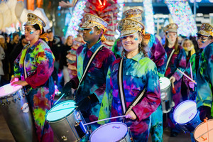 A group of people in colorful outfits and gold hats play drums in a festive parade, surrounded by bright lights and decorations at night.