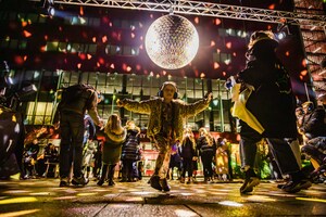 A child dances joyfully under a large disco ball, surrounded by people in a colorful, illuminated outdoor night scene.
