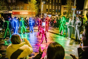 Performers in glowing LED suits dance at night in a city plaza, surrounded by a crowd of onlookers who are applauding. The illuminated suits create bright, colorful light against the urban backdrop.