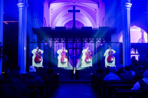 A church interior lit with purple lights features holograms of a cellist in white playing on stage, with an audience seated in pews watching the performance.