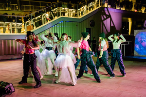 A group of young dancers perform outdoors at night, wearing glowing tutus or dark pants and beanies, as colorful lights illuminate the scene. Spectators watch from upper railings in the background.