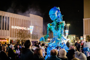 A large blue puppet resembling a person is paraded through a crowd at night. People watch the colorful spectacle, with illuminated buildings and a “SUPERBOWL UK” sign in the background.