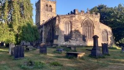 A stone church with arched windows and a square tower stands in a graveyard with old headstones and trees under a clear blue sky, illuminated by sunlight.