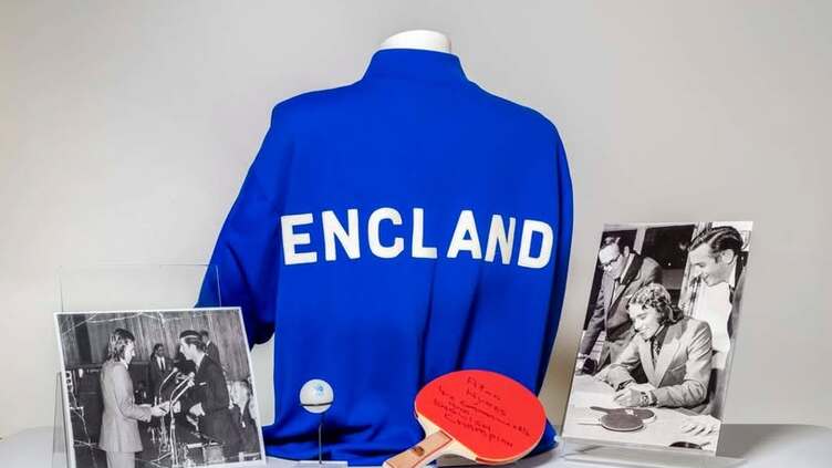 A blue jacket with "ENGLAND" on the back is displayed with a red table tennis paddle, a ping pong ball, and two black-and-white photos, one showing people signing papers and another showing a handshake.