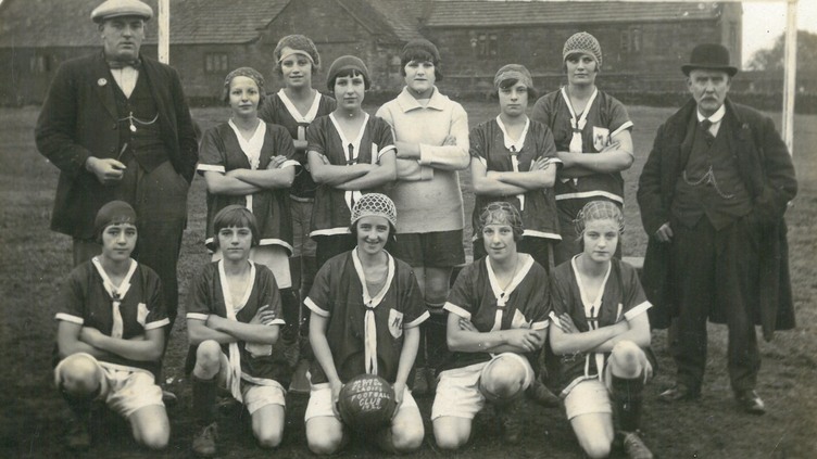 A vintage black-and-white photo of a women's soccer team and two men standing outdoors in front of a goalpost. The women wear uniforms and headbands, while the men are dressed in suits and hats.