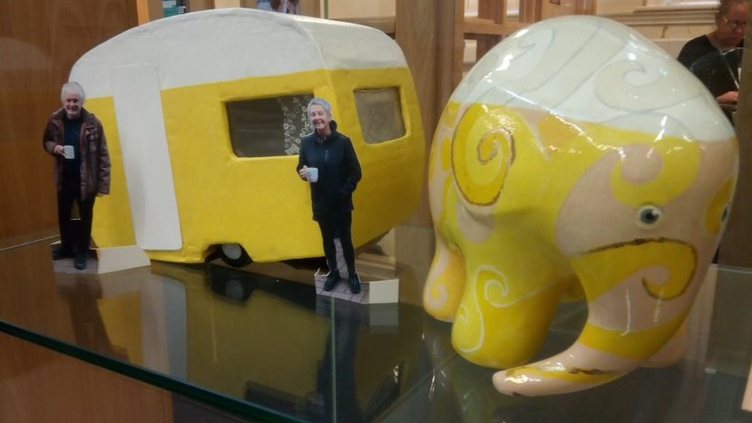 A small yellow and white ceramic elephant with a yellow caravan model behind it