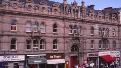 A row of shops including a hair salon, chemist, restaurant, and bakery sits beneath an ornate, historic stone building with round windows and archways. People walk along the sidewalk in front.