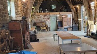 Interior of a rustic barn with stone walls, exposed beams, old machinery, a blue wheelbarrow, tables, and various vintage items arranged around the space. Light streams through small windows.