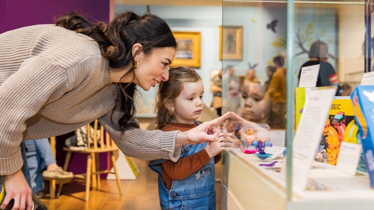 A woman and a young child look closely at items displayed in a glass museum case. The woman points at something inside, while the child watches with interest. The scene is colourful and warmly lit.