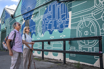 A man and a woman looking at a mural at Elsecar Heritage Centre. The mural is blue, green and white and features industrial images such as steam trains and wheels.
