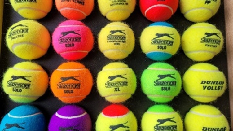 A collection of brightly colored tennis balls arranged in a grid. The balls display various brand names and logos, including Slazenger and Dunlop, and come in yellow, orange, red, green, purple, and blue.