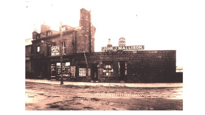 Sepia-toned photo of an old street with stone buildings, including shops with signs reading "John J. Mallison" and "Bain &amp; Luxford." The street appears wet, and there are chimneys on the buildings.
