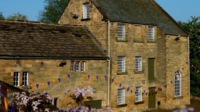 A stone building with barred windows, green doors, and a sloped roof is decorated with colorful triangular bunting. There are trees and greenery in the background under a clear blue sky.