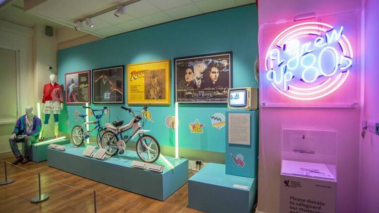 A view of the I grew Up 80s exhibition including a neon sign, mannequins in 80s dress, movie posters and two bicycles