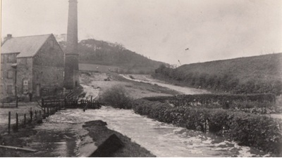 Black and white photo of a flooded rural scene with water flowing over a road near a large stone building and a tall chimney. Bushes and trees line the flooded area, and hills are visible in the background.