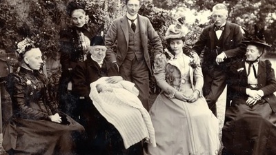 A vintage black-and-white photo shows seven adults in formal Victorian attire and one adult holding a baby wrapped in a blanket, posed outdoors with trees in the background.