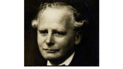 A black-and-white portrait of an older man with wavy light hair, a slight smile, and wearing a suit jacket and tie, looking directly at the camera.
