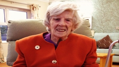 An elderly woman with short white hair sits in a cozy living room. She is wearing a bright orange coat with a pin, smiling, with a walking aid beside her. The background features a patterned wall and sofa with cushions.