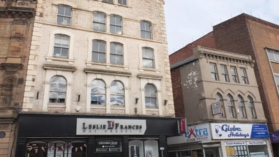 A row of commercial buildings features a white-brick hairdressing salon, “Leslie Frances,” with arched windows, next to shops “Xtra” and “Globe Holidays,” set beneath a cloudy sky.