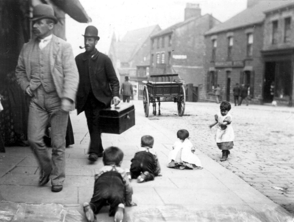 A black and white photograph of a street scene from the past. In the foreground, four children are sitting and playing on the sidewalk.
