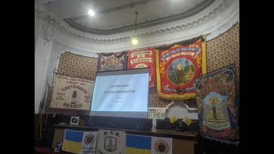 A presentation screen stands on a stage decorated with ornate miners’ banners and flags, including a Ukrainian flag, inside a traditional hall with patterned wallpaper and a curved ceiling.