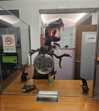 A black creature sculpture with red accents is on display behind a counter with a clear barrier. It is next to a fan, a no smoking sign, and some small objects. A "Staff Only" door is visible in the background.