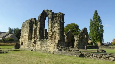 The image shows the stone ruins of an old building with tall arched windows, surrounded by grass. Trees and houses are visible in the background under a clear, blue sky.