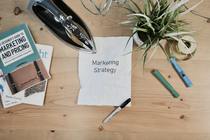 A desk with stationary and a pot plant and a piece of paper saying 'marketing strategy'