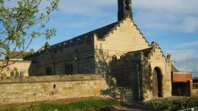 A small stone church with a stepped roofline and bellcote, surrounded by a low stone wall and greenery, under a blue sky with scattered clouds.