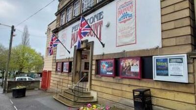 A historic theater building with posters and Union Jack flags on the exterior, steps leading to the entrance, a black trash bin, and a flower planter in the foreground. Trees and a parked car are visible to the left.