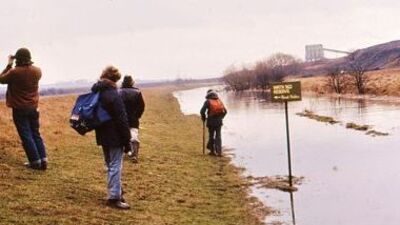 Four people with backpacks walk along a grassy path beside a flooded river. One person takes a photo, while a sign partially submerged in water reads "DO NOT WALK" with an industrial building visible in the distance.