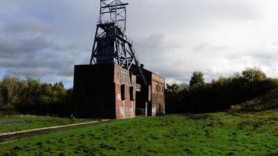 A brick industrial building with a tall metal mining tower stands on a grassy field, surrounded by trees under a cloudy sky.