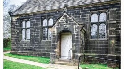 A historic stone building with arched windows, a pitched roof, and a central entrance with a white door. A paved path curves in front, with green grass and trees in the background.