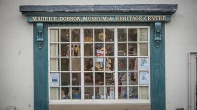 A large window with blue-green trim displays various items inside the Maurice Dobson Museum &amp; Heritage Centre, which is identified by a sign above the window. Posters and decorations are visible behind the glass.