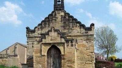 Small historic stone chapel with arched doorway and bell tower, featuring scalloped edges on the roof. The building stands under a blue sky with a tree and houses visible in the background.