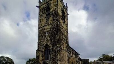 Stone church with a tall square tower, arched windows, and spires at the corners, set against a cloudy sky with grass and trees surrounding the building.