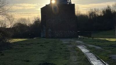 A narrow path leads to an old brick building with a metal tower, silhouetted by the setting sun, surrounded by grass and bare trees.