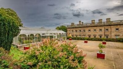 A large garden with a white glass conservatory, red planters on a paved area, lush greenery, and a historic stone building under a cloudy sky.