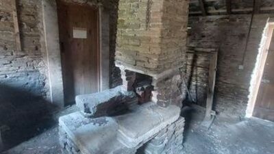 An old, crumbling brick fireplace stands in a dusty, abandoned room with exposed brick walls, wooden doors, and debris scattered on the floor.