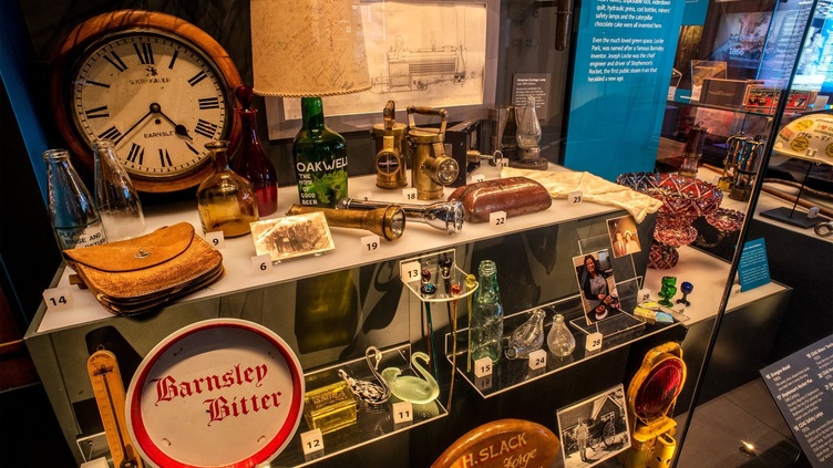 A display case in Experience Barnsley with a number of objects including a clock, lampshade made from a beer bottle, glass swans and bottles and miner's lamps.