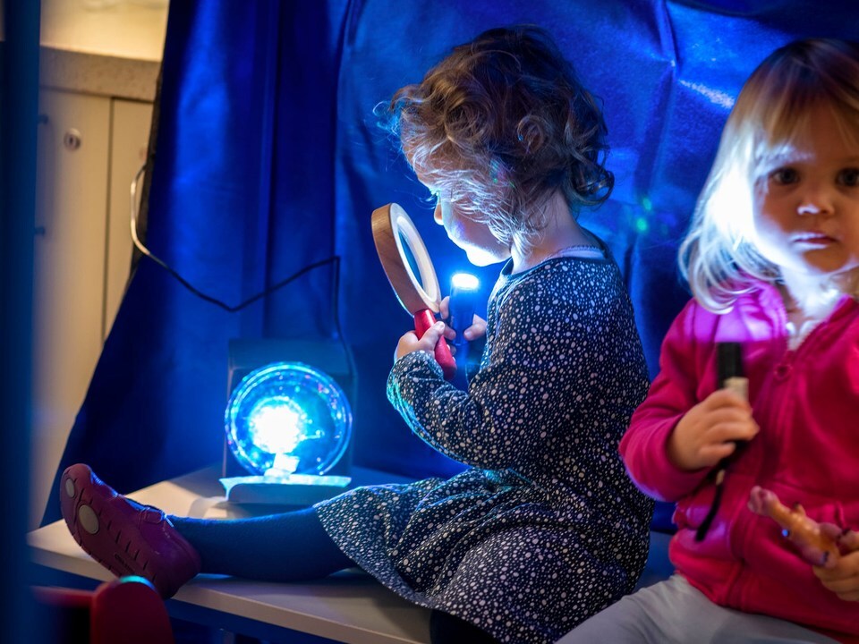 Two young children explore light and reflection. One child holds a magnifying glass under a blue light, while the other shines a flashlight.