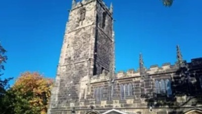 A tall stone church tower with a clock, set against a clear blue sky, surrounded by trees with autumn foliage.